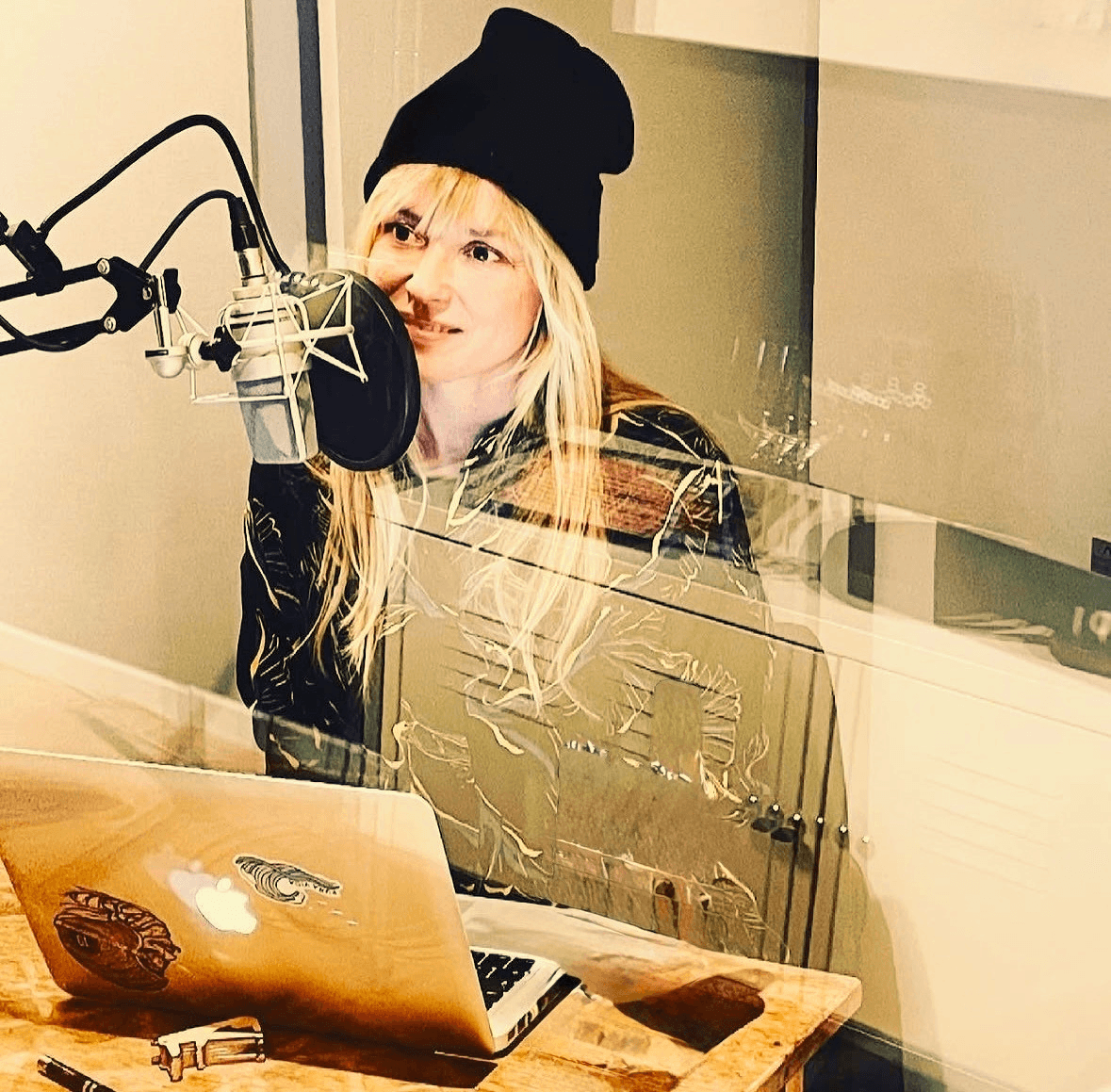 Kerstin Kleinenbrands mit Beanie am Studiomikrofon, hinter der Glasscheibe eines Aufnahmeraums